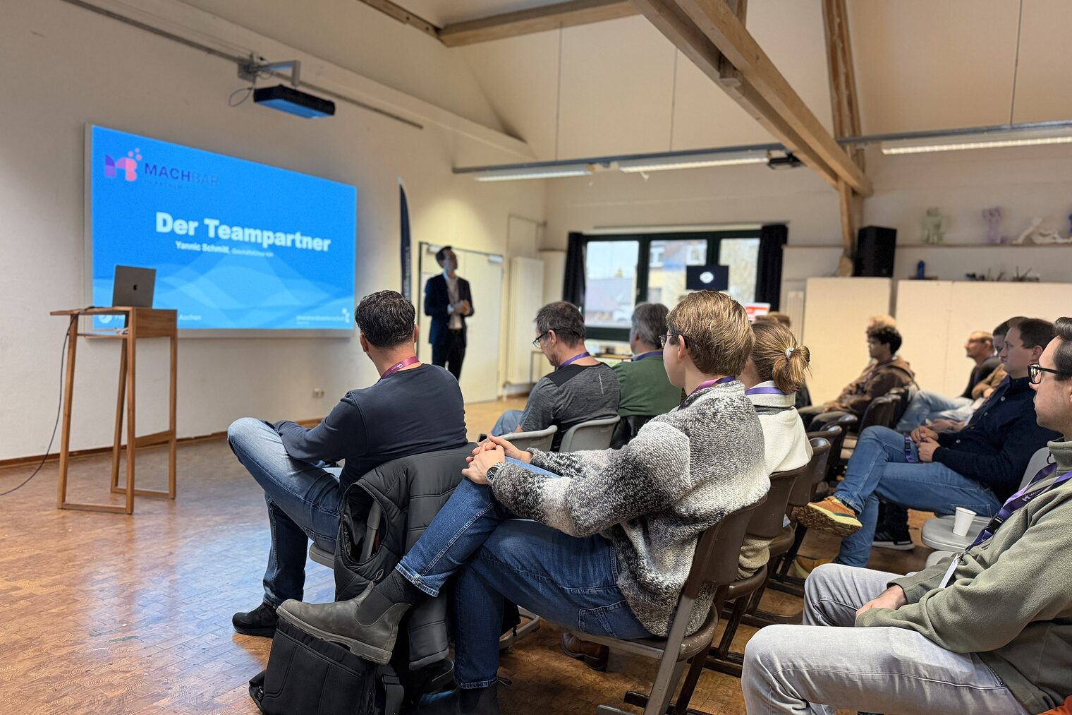 Eine Gruppe von Menschen sitzen vor einem Redner. Im Hintergrund sieht man eine Präsentation mir der Folie "Der Teampartner"