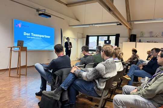 Eine Gruppe von Menschen sitzen vor einem Redner. Im Hintergrund sieht man eine Präsentation mir der Folie "Der Teampartner"