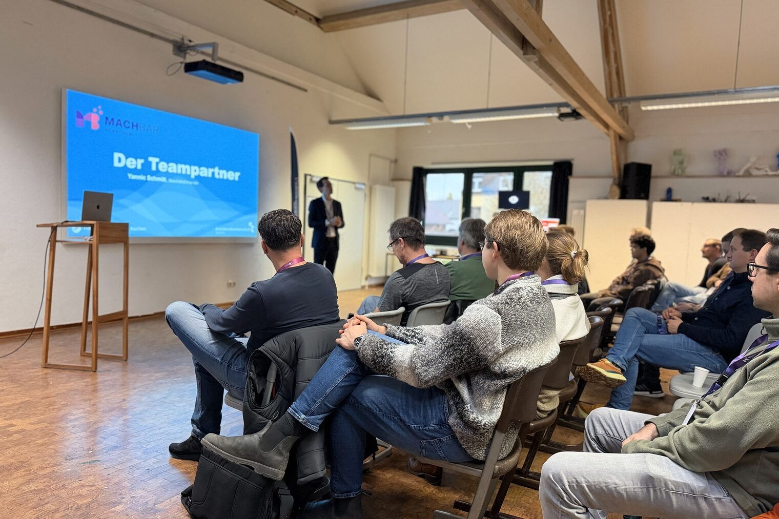 Eine Gruppe von Menschen sitzen vor einem Redner. Im Hintergrund sieht man eine Präsentation mir der Folie "Der Teampartner" 