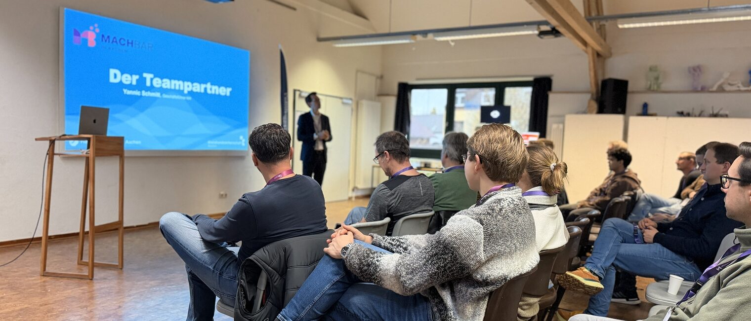 Eine Gruppe von Menschen sitzen vor einem Redner. Im Hintergrund sieht man eine Präsentation mir der Folie "Der Teampartner" 